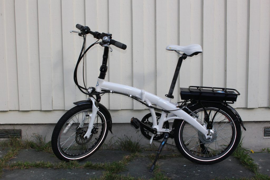 En elcykel står framför en vit vägg. Den har små hjul och en vit ram som är ganska bred. Under pakethållaren syns en liten svart låda.