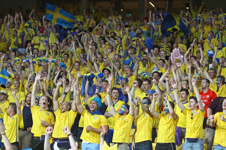 Svenska fotbollssupportrar står på en läktare och hejar på det svenska landslaget.