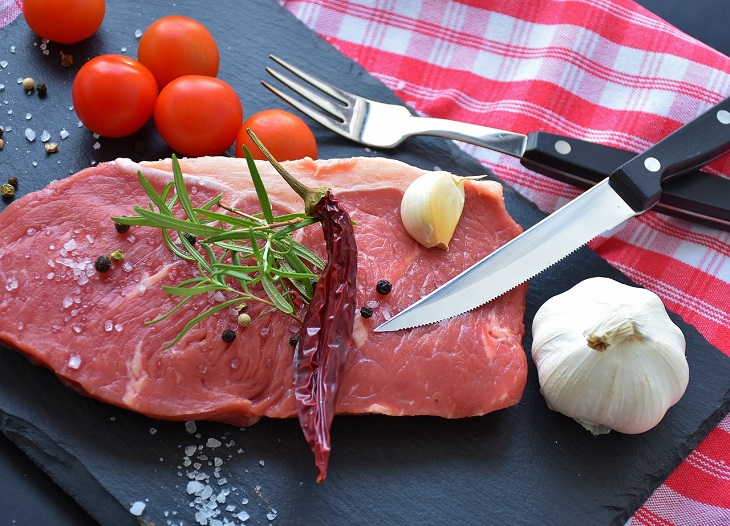 En rå köttbit ligger på en skärbräda. På köttbiten ligger en kryddkvist och en torkad chili. Bredvid köttebiten ligger en kniv, en vitlök och några tomater. Det är strött peppar och salt över köttbiten och skärbrädan.