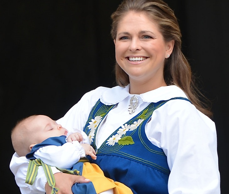 Prinsessan Madeleine har svensk folkdräkt på sig som är gul och blå. Hon ler stort och håller en bebis i sina armar.
