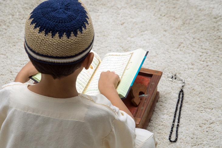 Ett barn sitter med ryggen mot oss. Framför sig har det en uppslagen koran på en bricka. Bredvid ligger ett ban med pärlor. Barnet har en virkad lite mössa på sig.