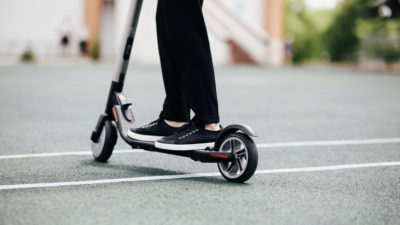 Nedre delen av en elsparkcykel där man ser en persons ben stå på ståplattan. Den åker fram över en asfaltsväg.